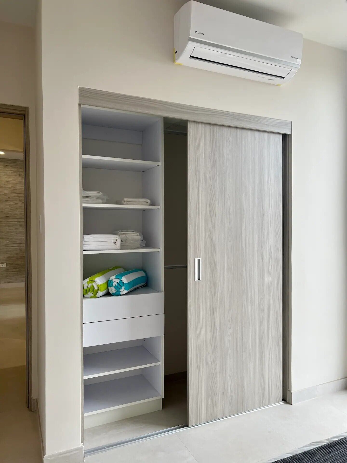 Ein begehbarer Kleiderschrank mit Regalen und einer Schiebetür, eine Klimaanlage darüber und ein gefliester Boden.