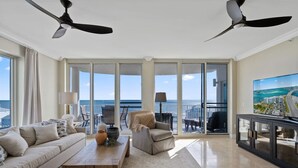 A spacious living room with a large sofa, a coffee table, and a TV on a stand. There are two ceiling fans, a balcony with a view of the ocean, and a large window. - Mustique 802- Luxury Condo with Direct Gulf View a (Gulf Shores)