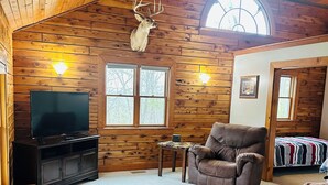 A cozy cabin interior with a wooden ceiling, a mounted deer head, a television, a leather sofa, and a window with a view of trees.
