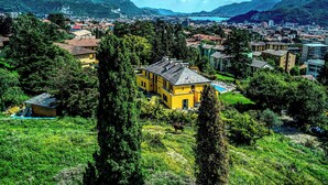 A yellow building surrounded by greenery, with a mountainous backdrop and a lake in the distance. - Montebello Lake (Lecco)