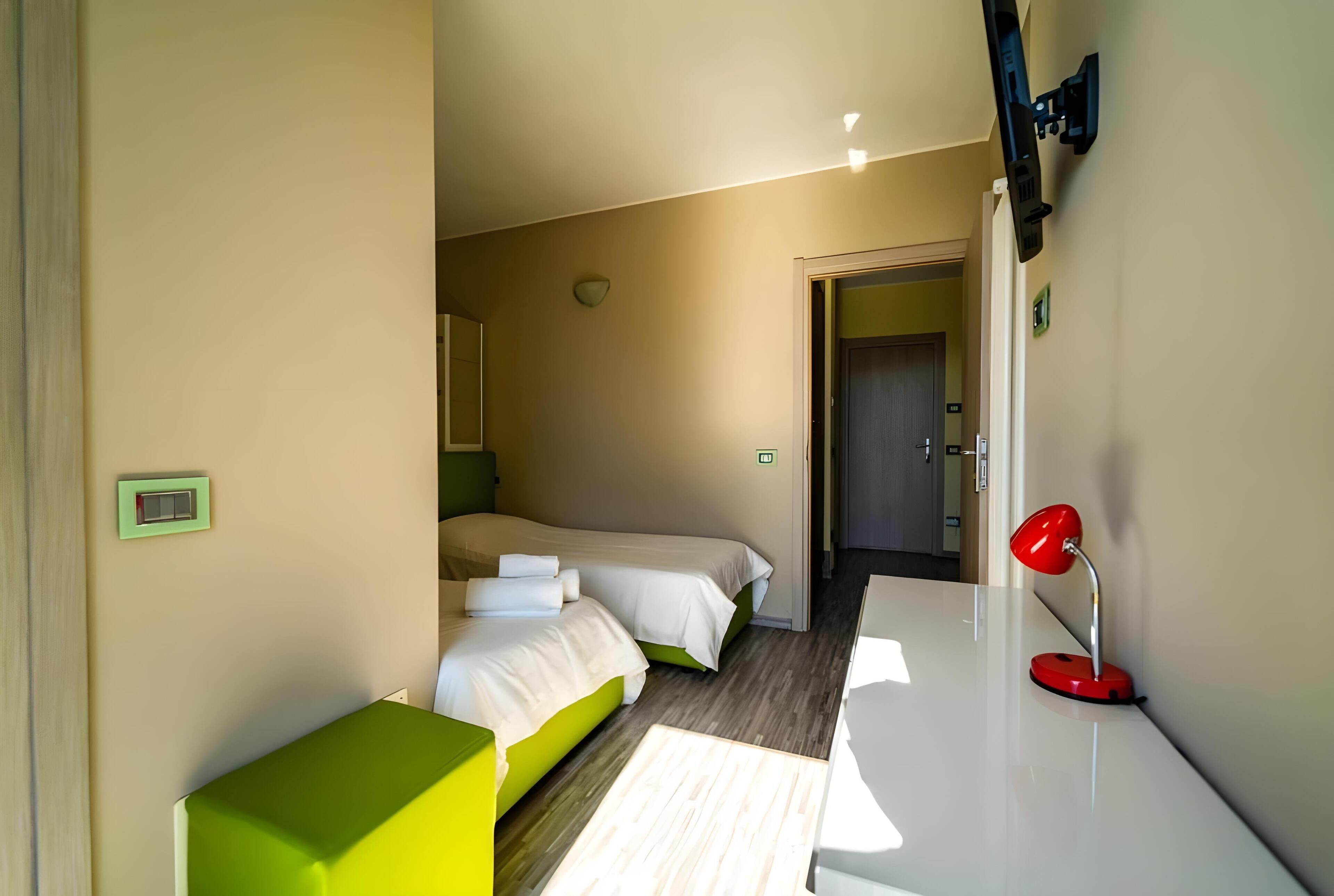 Une chambre d’hôtel avec deux lits, un bureau, une lampe de bureau rouge et un téléviseur fixé au mur.