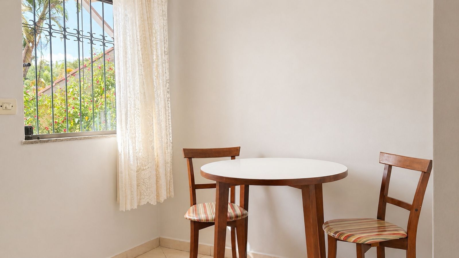 Una habitación pequeña con una mesa redonda y dos sillas de madera, una ventana con cortina blanca y vistas al exterior con vegetación.