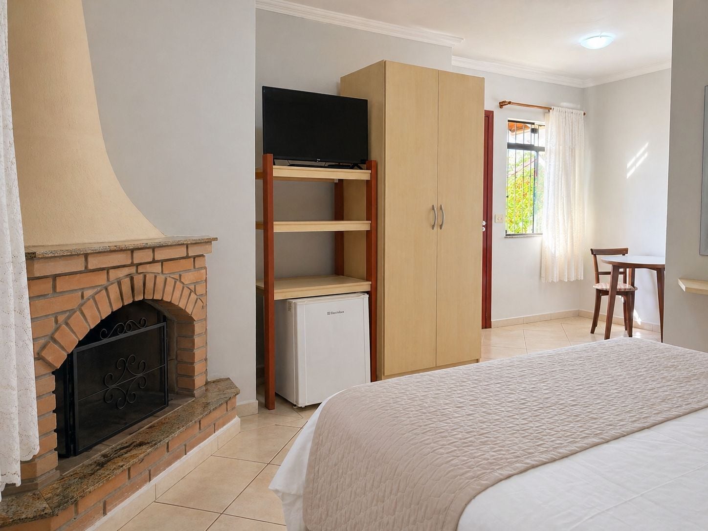Un dormitorio con chimenea de ladrillo, un televisor en un estante, un armario, una mesita y una ventana con cortinas.