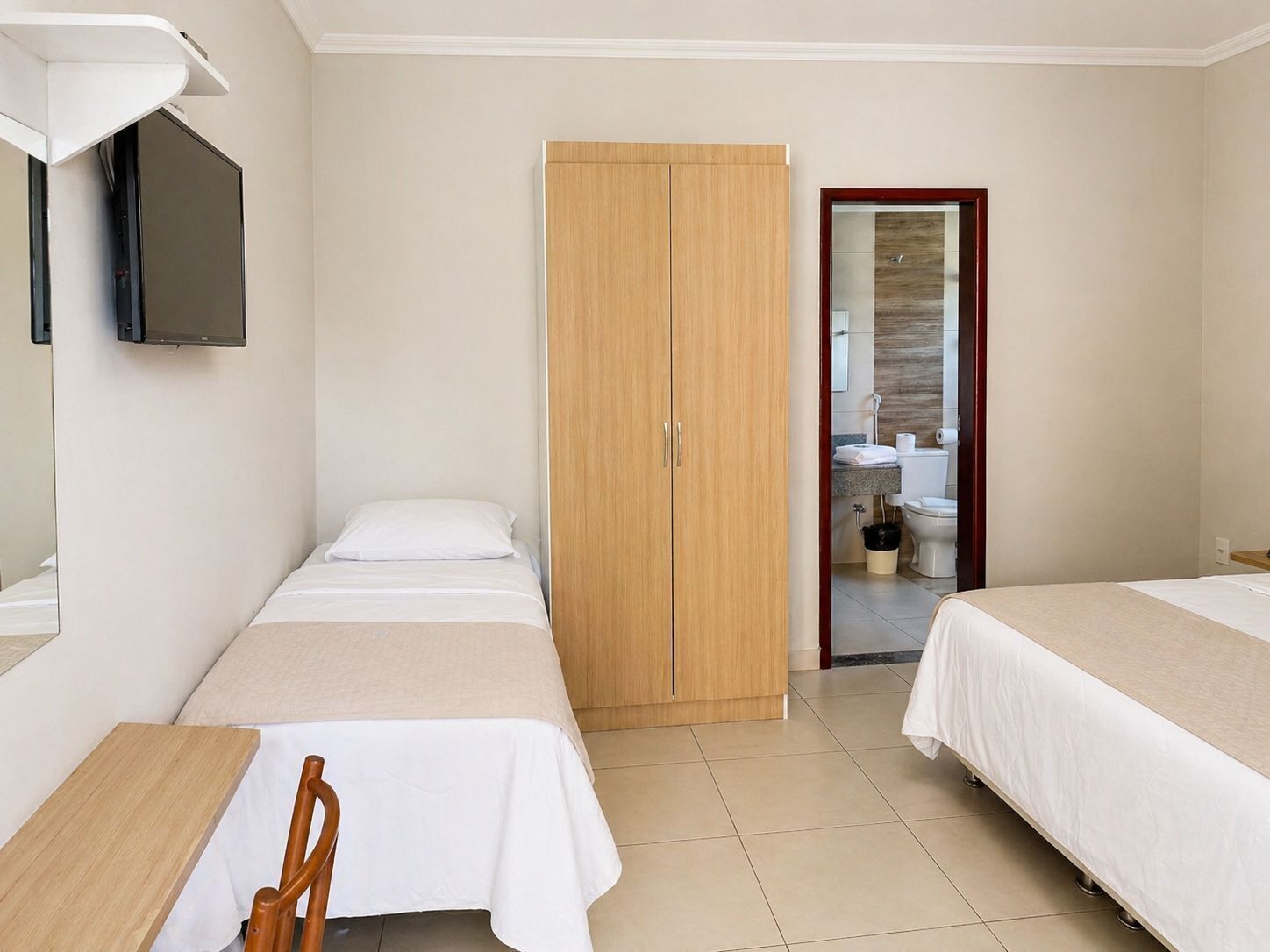 Quarto de hotel com duas camas, guarda-roupa de madeira, televisão na parede e banheiro visível por uma porta aberta.