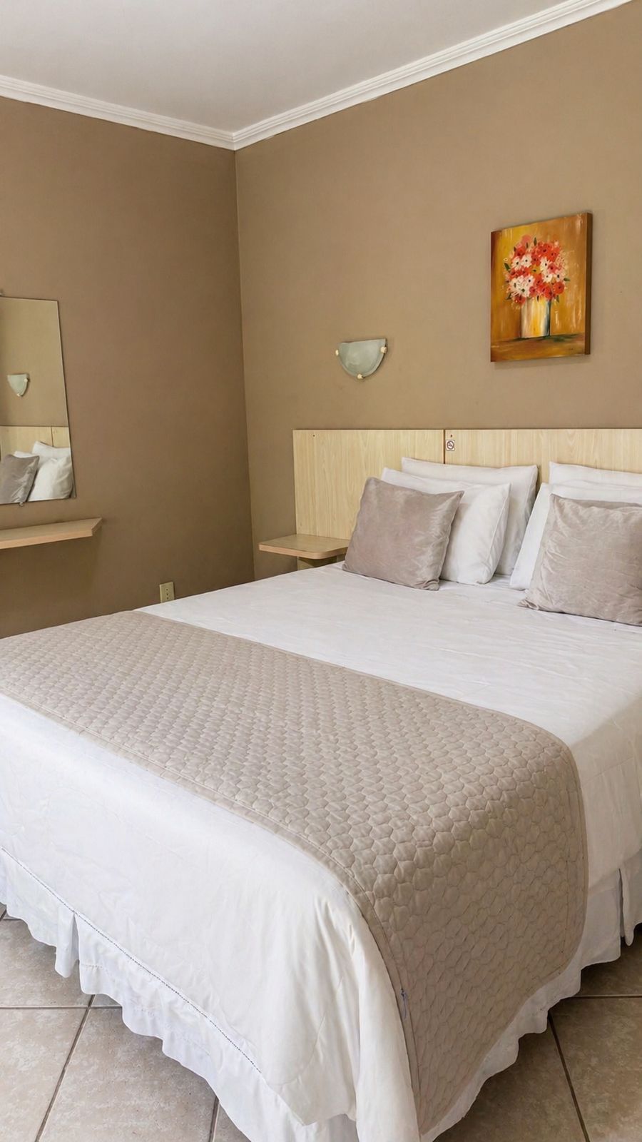 Quarto de hotel com uma cama grande, cabeceira, mesa de cabeceira, espelho fixado na parede e uma pintura.
