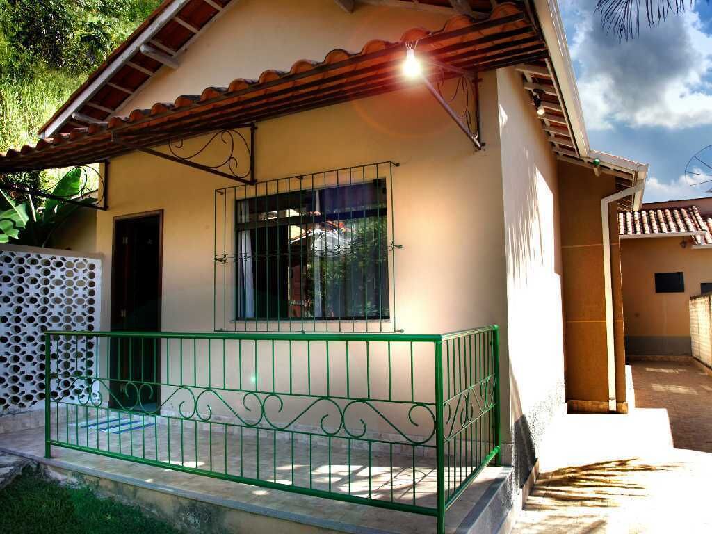 Uma casa térrea com telhado de telhas, varanda com grades verdes e uma janela com barras.