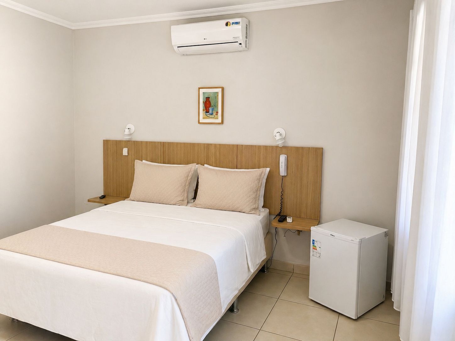 A hotel room with a bed, bedside tables, a small refrigerator, an air conditioner, a painting, and a telephone.