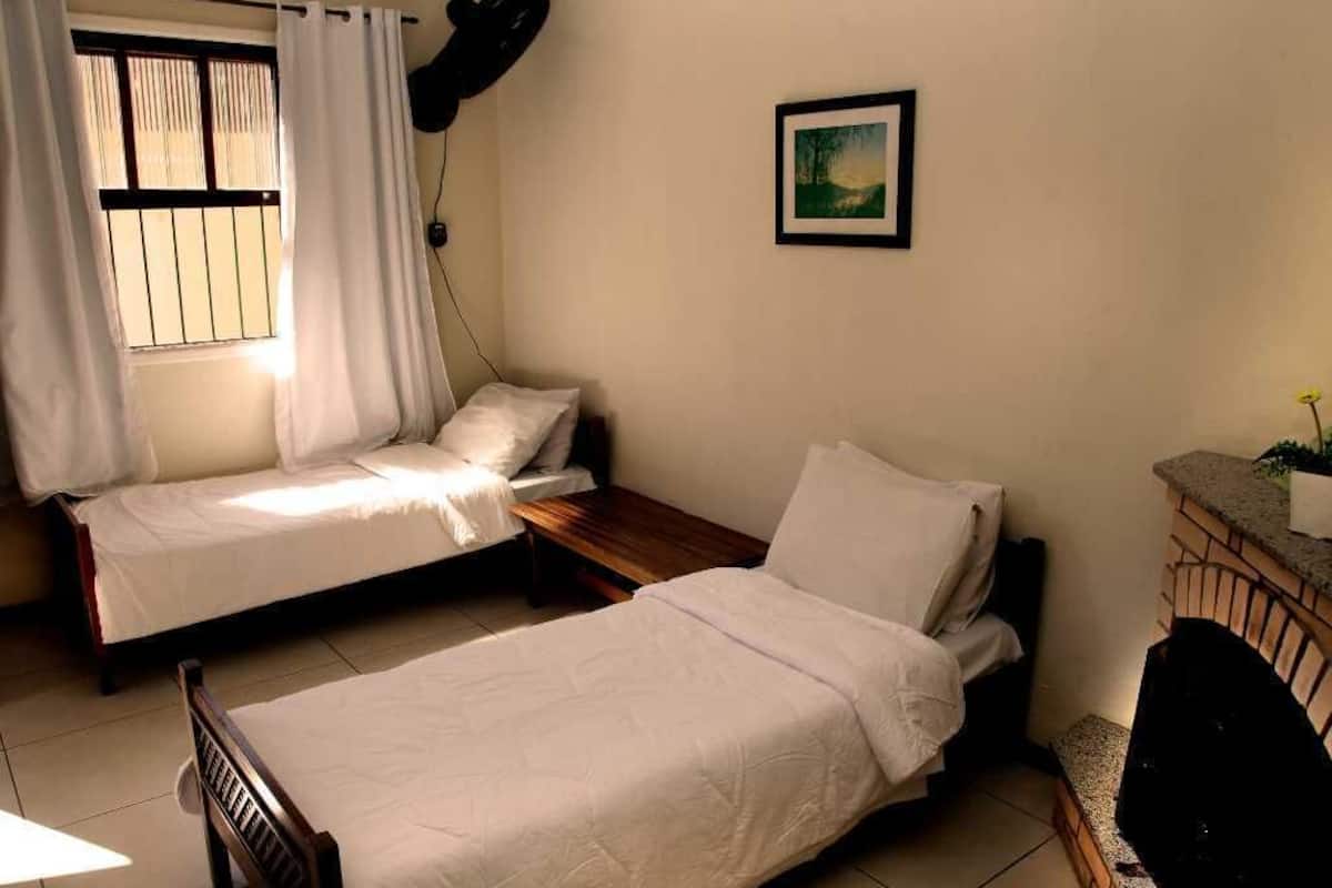 Ein Zimmer mit zwei Einzelbetten, einem kleinen Tisch, einem Stuhl, einem Fenster mit Vorhängen, einem gerahmten Bild an der Wand und einer Ziegel-Theke.