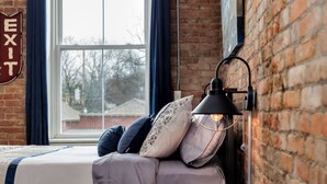 Et soveværelse med en murstensvæg, en seng med et blåt og hvidt stribet sengetæppe, flere puder, et rundt natbord og en sort lampe.