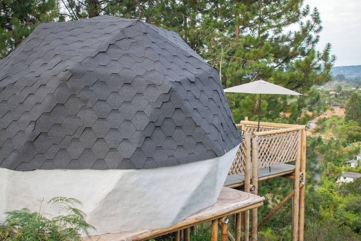 A geodesic dome structure with a white base, surrounded by a bamboo deck and an umbrella.