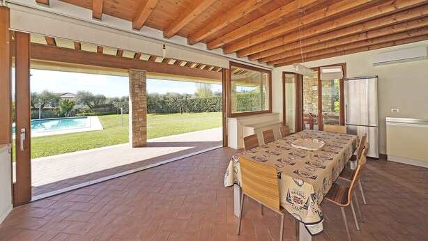 An open-plan living area with a dining table, wooden ceiling beams, large windows, and a view of a swimming pool.