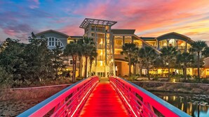 A hotel with a red bridge leading to a building with multiple levels and balconies, surrounded by palm trees and a body of water. - Golf Cart, 30A Watersound Gated Community, Beach Views, Gulf Front, Large Pool (Inlet Beach)