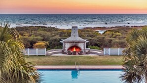 A pool with a stone fire pit and outdoor seating, surrounded by greenery and a view of the ocean at sunset. - Golf Cart, 30A Watersound Gated Community, Beach Views, Gulf Front, Large Pool (Inlet Beach)