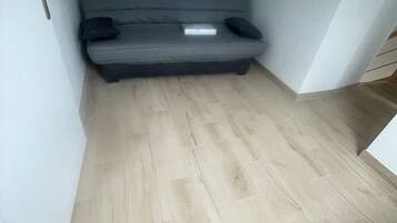 A room with a grey sofa bed against the wall, a wooden floor, and a door on the left.