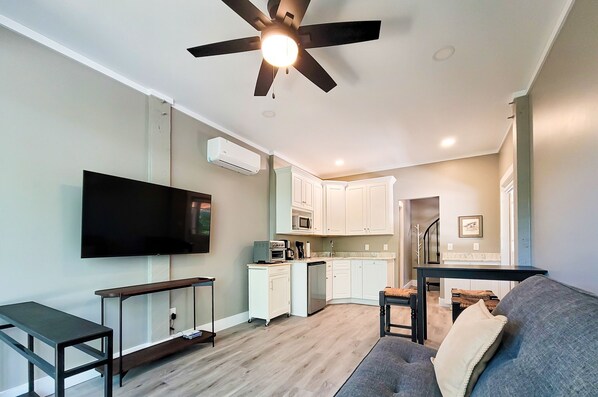 A modern living room with a flat-screen TV, a ceiling fan, a dining table, and a kitchen area with white cabinets and a microwave. - Beachcomber – Small Space, Big Paradise! (Topsail Beach)