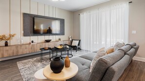 A modern living room with a large flat-screen TV mounted on the wall, a grey sectional sofa, a round coffee table with decorative vases, and a minimalist dining area with a black table and chairs. - Reunion Retreat 4-Bedroom Home w/ Playful Touches (Kissimmee)
