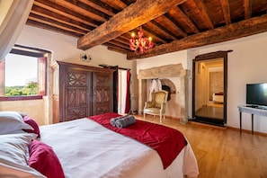 A spacious bedroom with a large bed, wooden ceiling beams, a chandelier, a television, and a view of the outdoors.