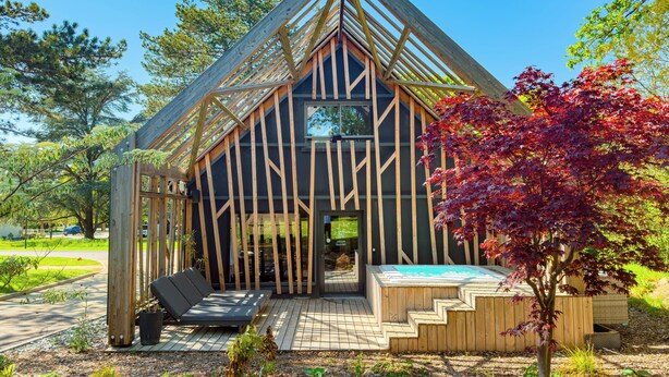 Una cabaña moderna de madera con techo inclinado, un patio con asientos y un jacuzzi pequeño.
