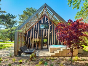 Una cabaña moderna de madera con techo inclinado, un patio con asientos y un jacuzzi pequeño.