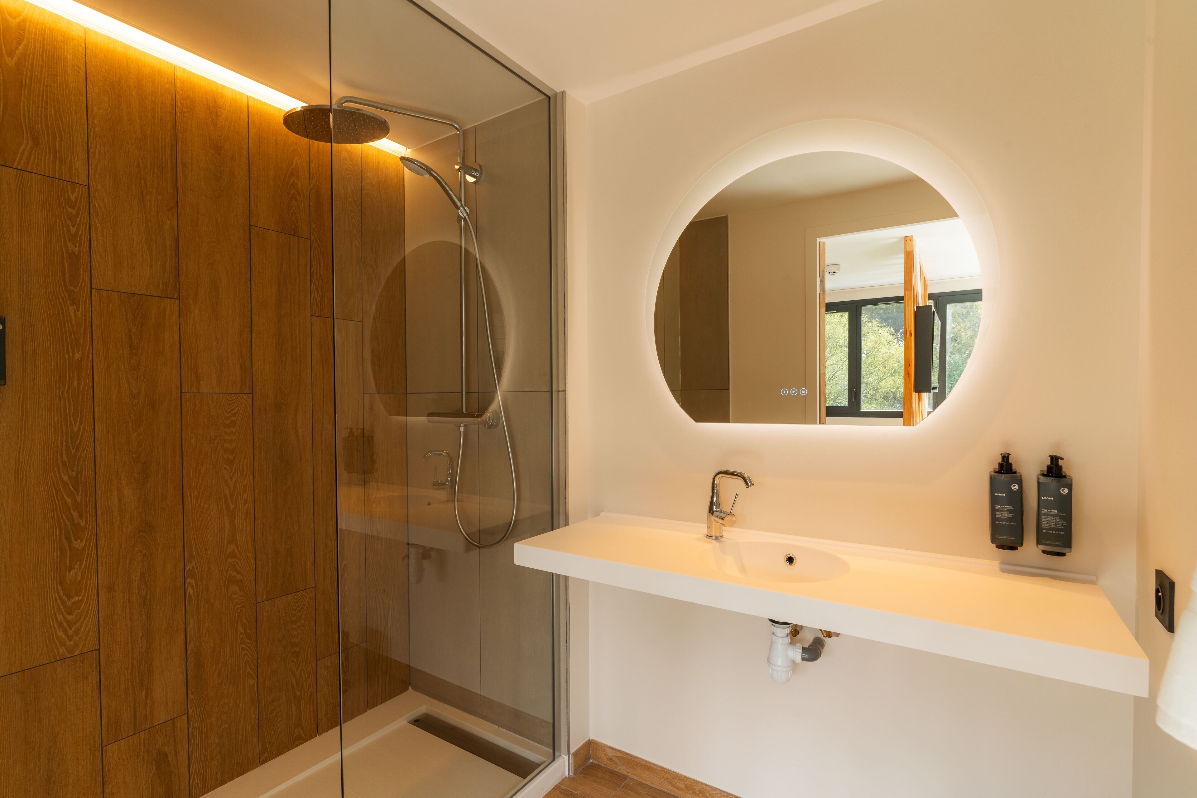 A modern bathroom with a glass-enclosed shower, a white sink with a round mirror, and wooden paneling on the wall.