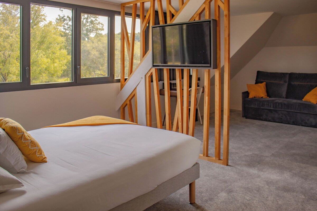 Un dormitorio con cama, televisor montado en un panel de madera, sofá con cojines y ventanas amplias con vista a árboles.
