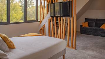 A bedroom with a bed, a TV mounted on a wooden partition, a sofa with cushions, and large windows with a view of trees.