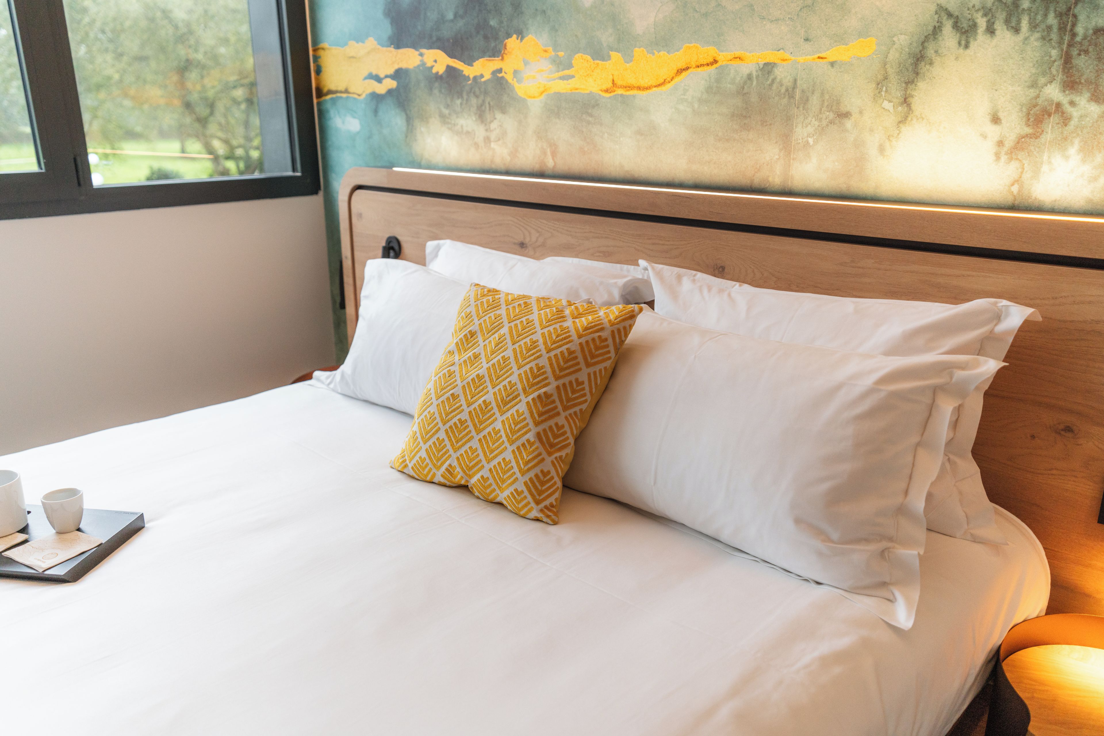 A bed with white bedding and a single patterned pillow, a bedside table with cups, and a view of a window with a painting above.