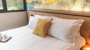 A bed with white bedding and a single patterned pillow, a bedside table with cups, and a view of a window with a painting above.