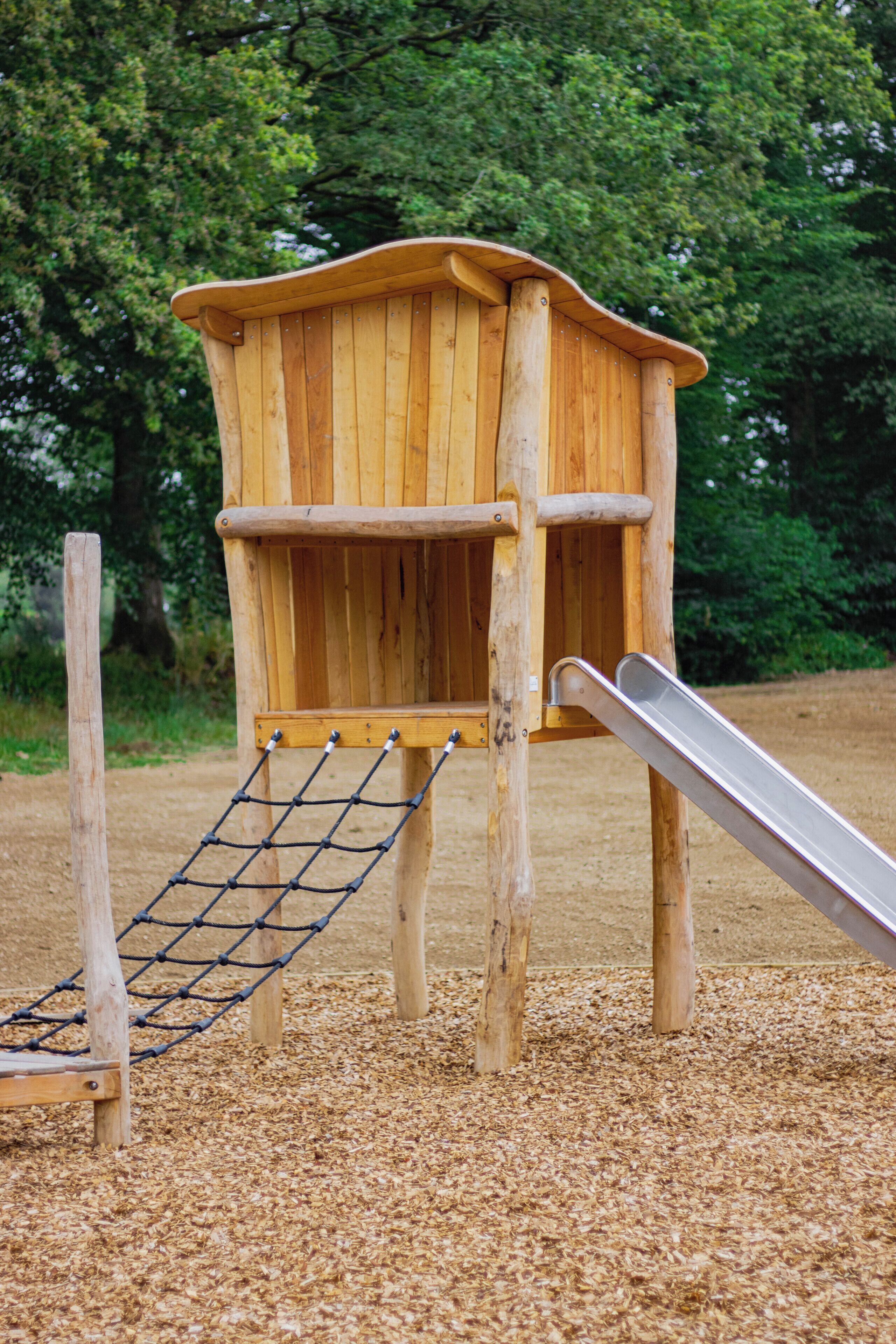 Une structure de jeu en bois, dotée d’un toboggan et d’un filet d’escalade, entourée de paillis.