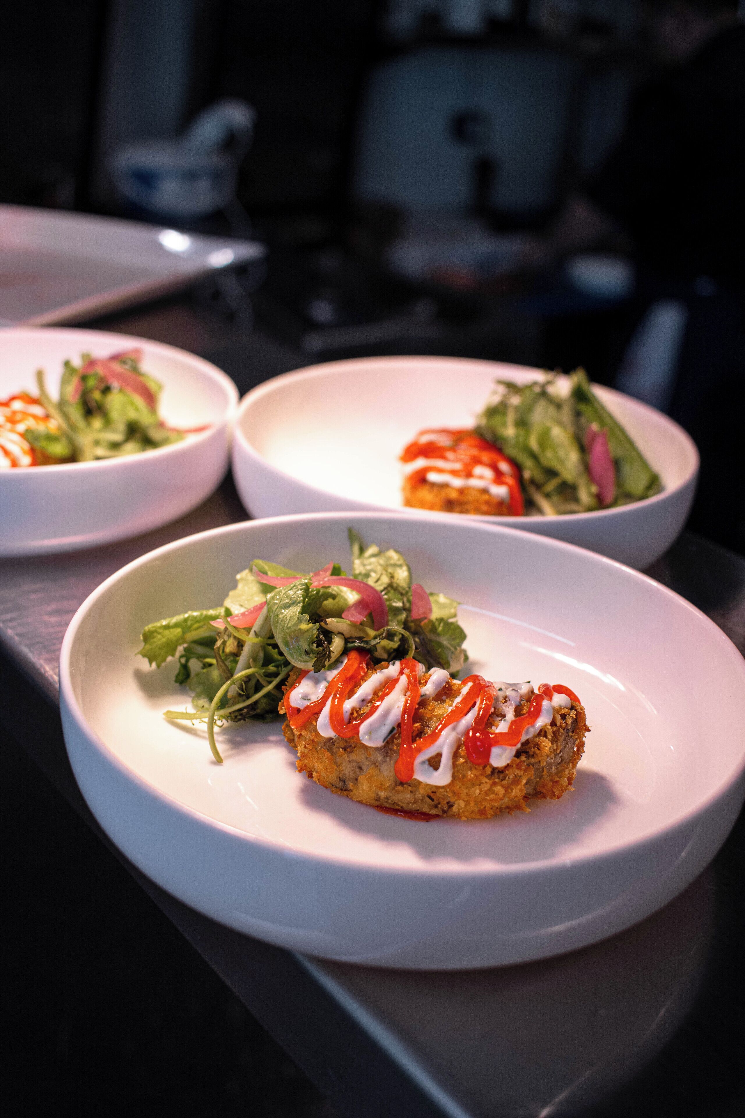 Tres platos blancos con comida, uno con un ingrediente empanizado y salsa, y dos con ensaladas.