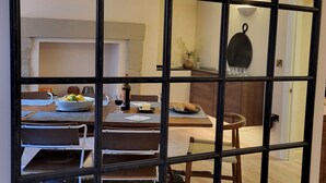 Un coin repas avec une table dressée pour deux personnes, un vase rempli de fleurs et une cuisine visible à travers une fenêtre à carreaux.