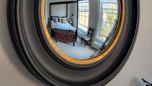 Un miroir rond, doté d’un cadre doré, reflète une chambre à coucher avec un lit, une chaise, un bureau et une fenêtre.
