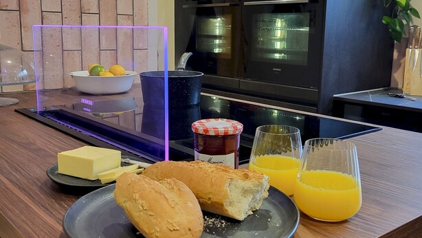 Un plan de travail de cuisine avec une assiette contenant du pain, du beurre, de la confiture et du jus d’orange.