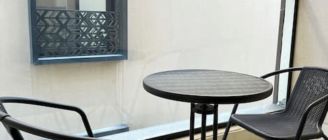 Un balcon avec une table ronde et deux chaises, une fenêtre ornée d’un motif géométrique et une enseigne indiquant « Kease ».