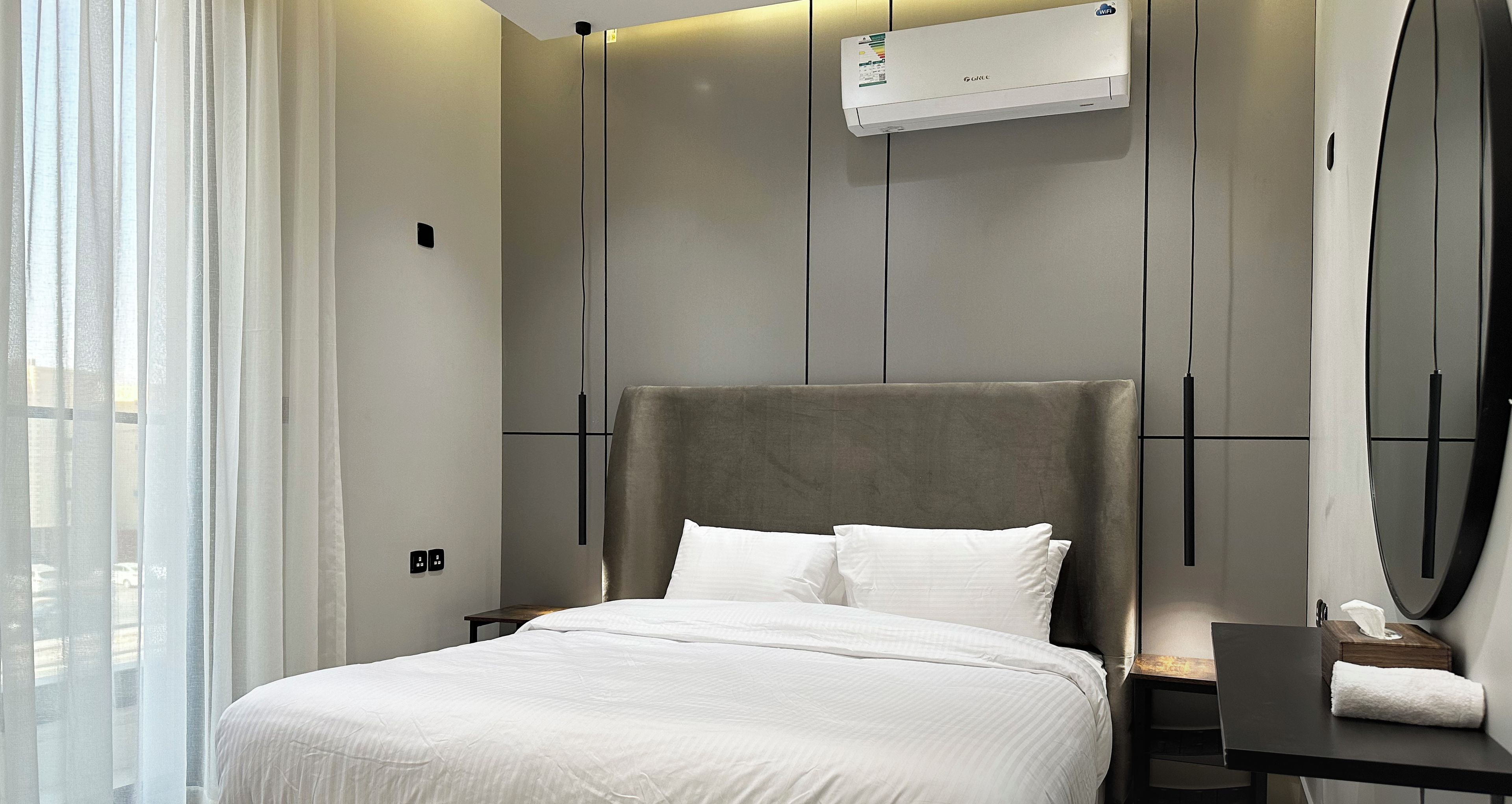 A modern hotel room with a large bed, a bedside table, a mirror, and an air conditioning unit.