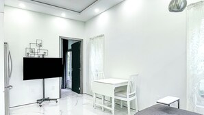 Une pièce moderne et bien éclairée, avec une table à manger blanche, des chaises, un canapé gris et un téléviseur à écran plat fixé au mur.