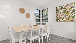 Un coin repas avec une table et des chaises blanches, une fenêtre avec des stores et une œuvre d’art accrochée au mur.