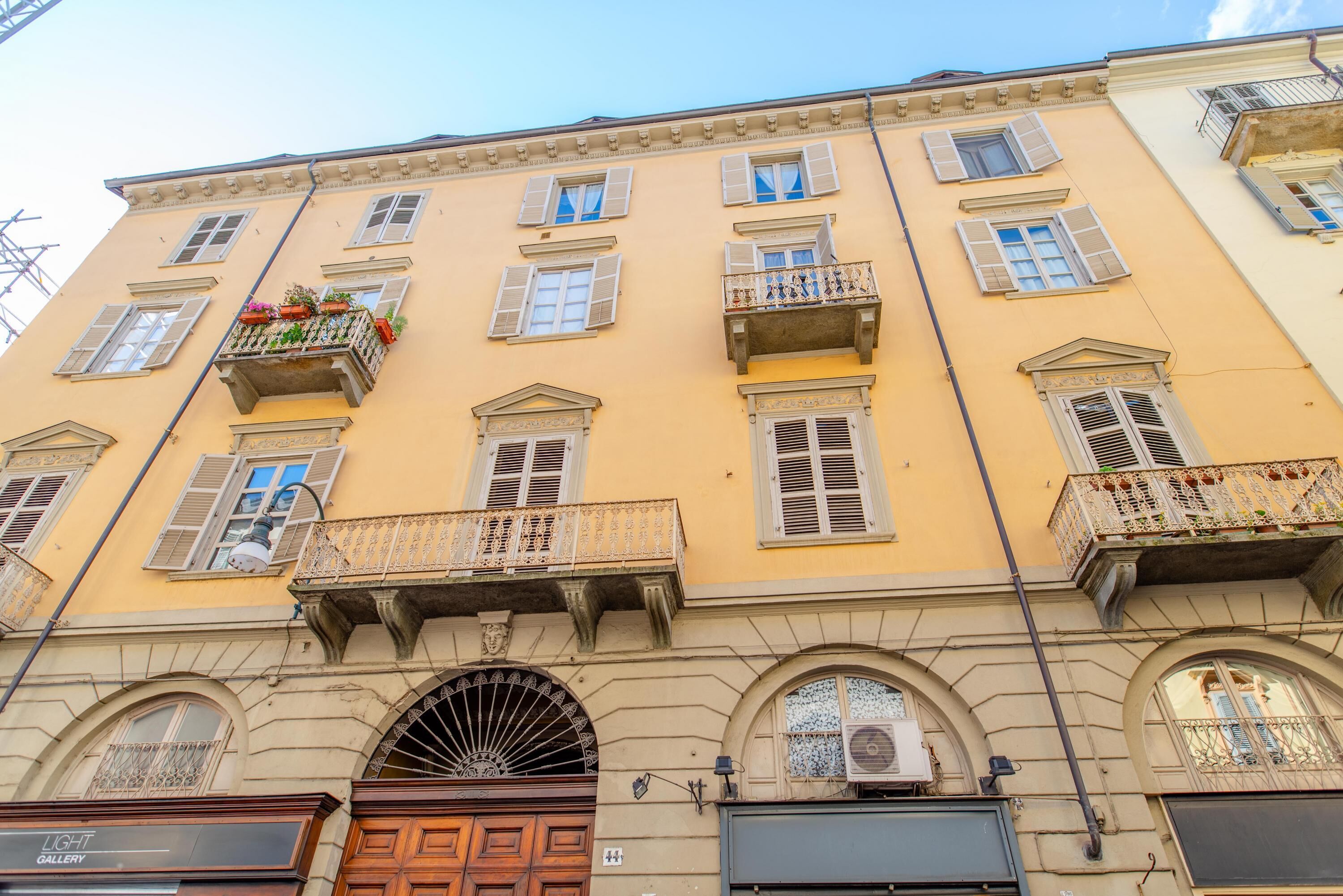 A multi-story building with a yellow facade, multiple balconies with flower boxes, and a large arched entrance.