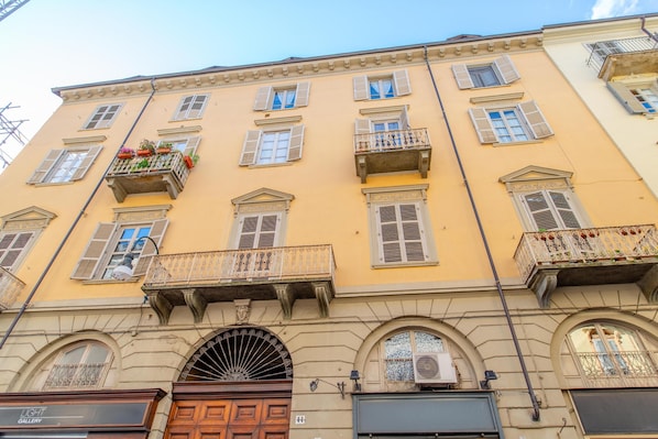 A multi-story building with a yellow facade, multiple balconies with flower boxes, and a large arched entrance.
