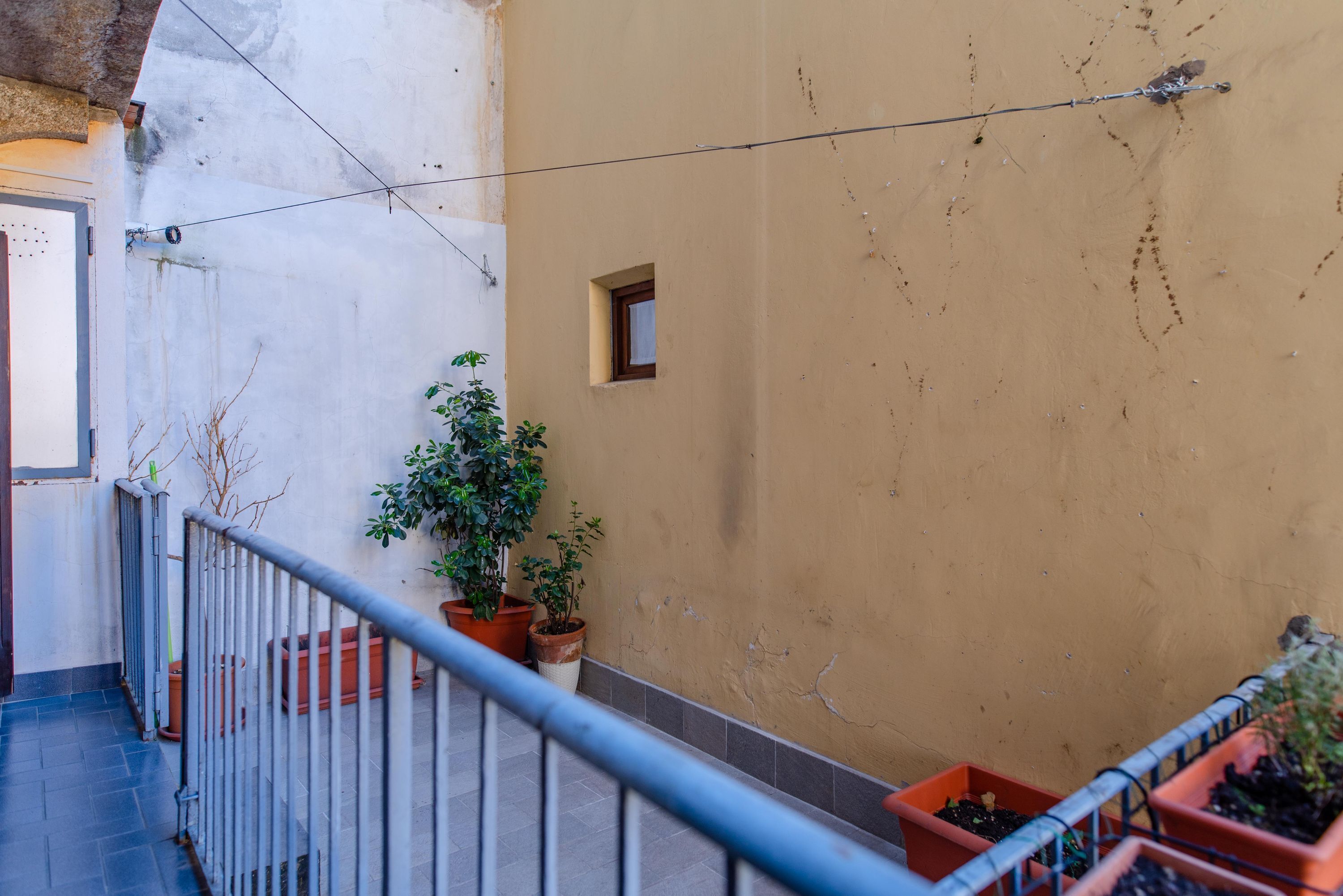 A narrow outdoor corridor with a metal railing, potted plants, and a small window.