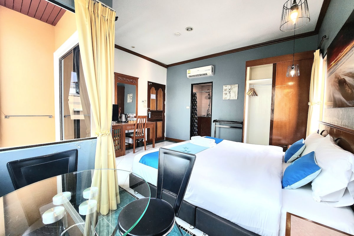 A hotel room with a bed, a desk, a chair, a glass table, and a bathroom visible through an open door.