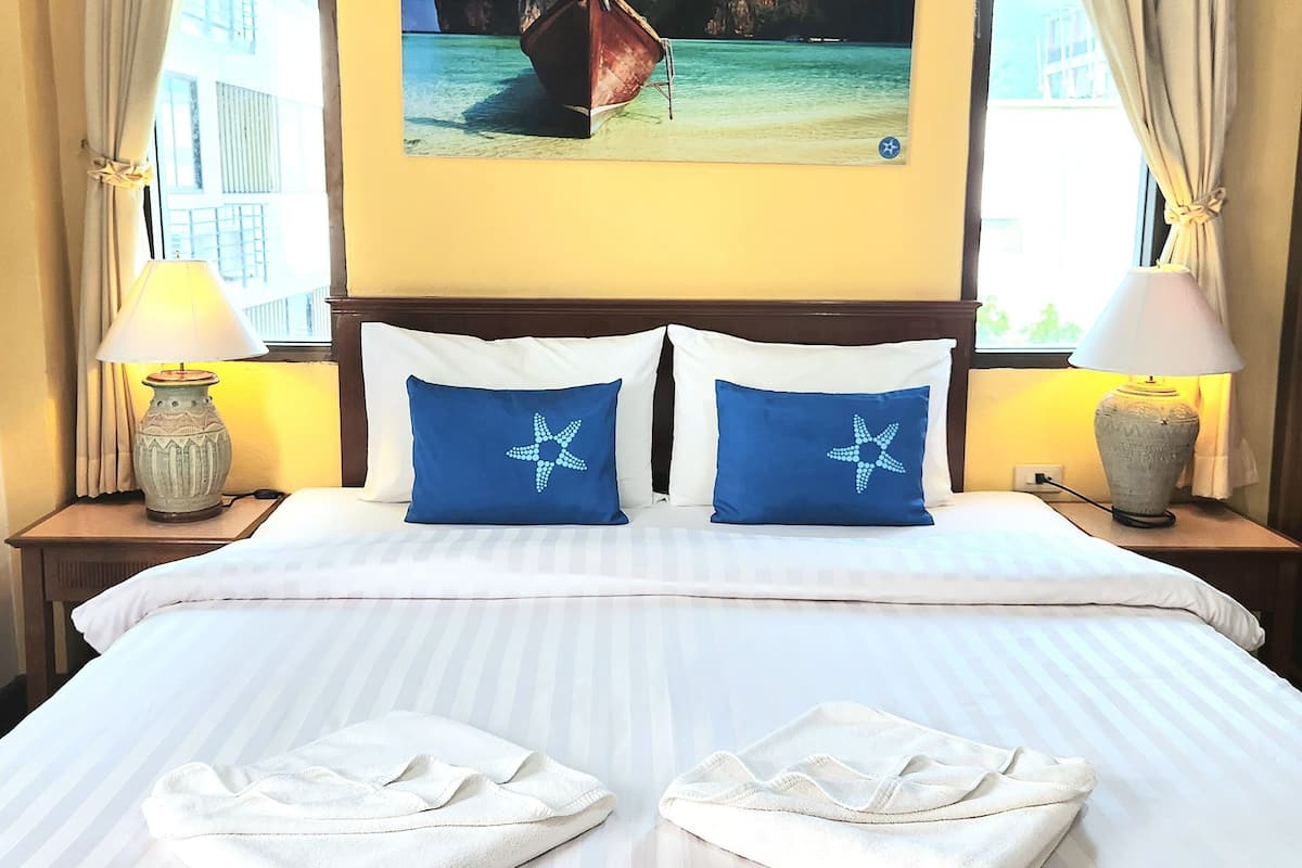 A hotel room with a bed, two nightstands, a painting of a boat on a beach, and curtains.