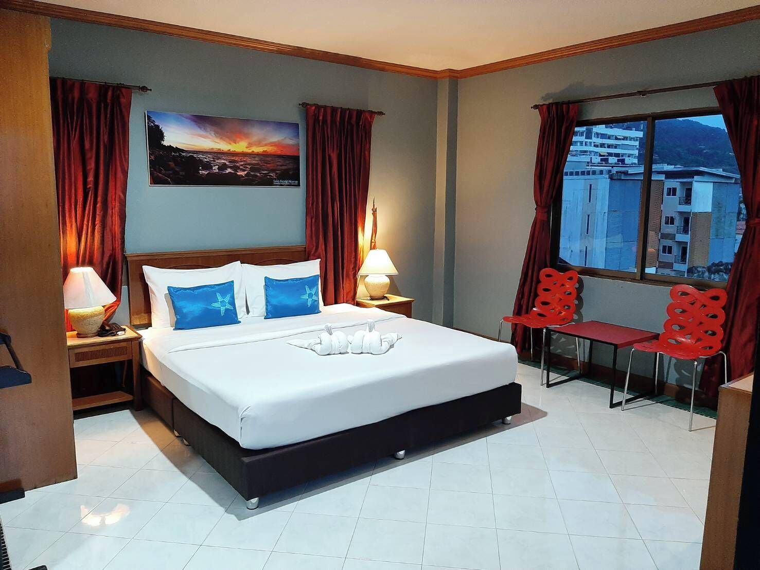 Habitación de hotel con cama, dos lámparas de noche, ventana con cortinas y vistas a edificios, además de una pequeña mesa con sillas.