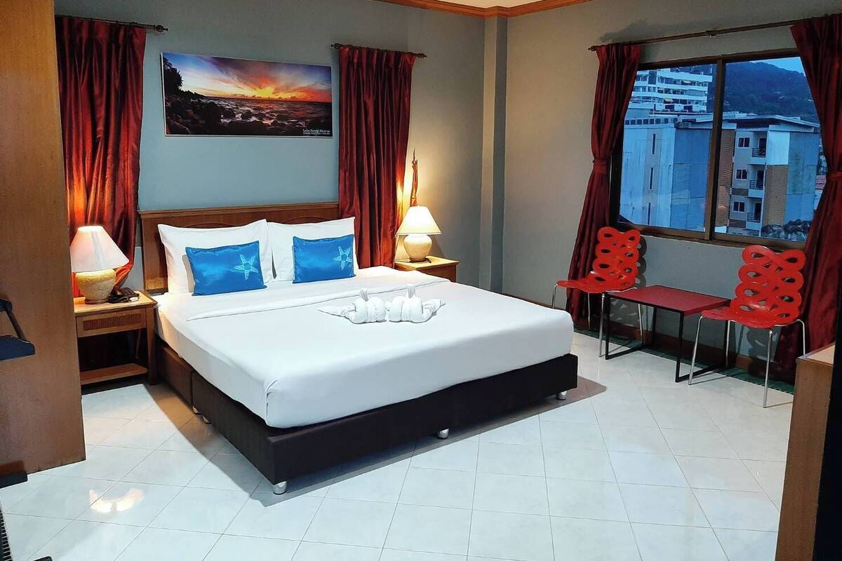 A hotel room with a bed, two bedside lamps, a window with a view of buildings, and a red chair by the window.
