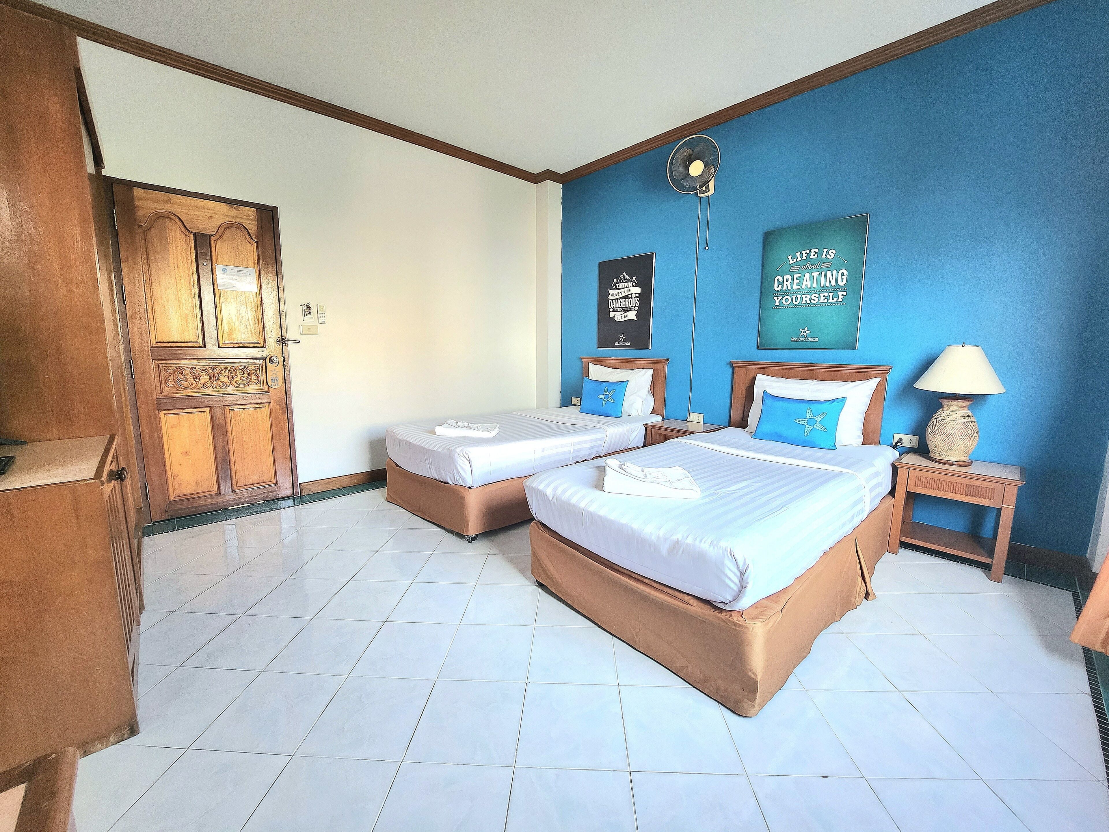 Habitación de hotel con dos camas, un armario de madera, una pared azul decorada con carteles motivacionales y suelo de baldosas.