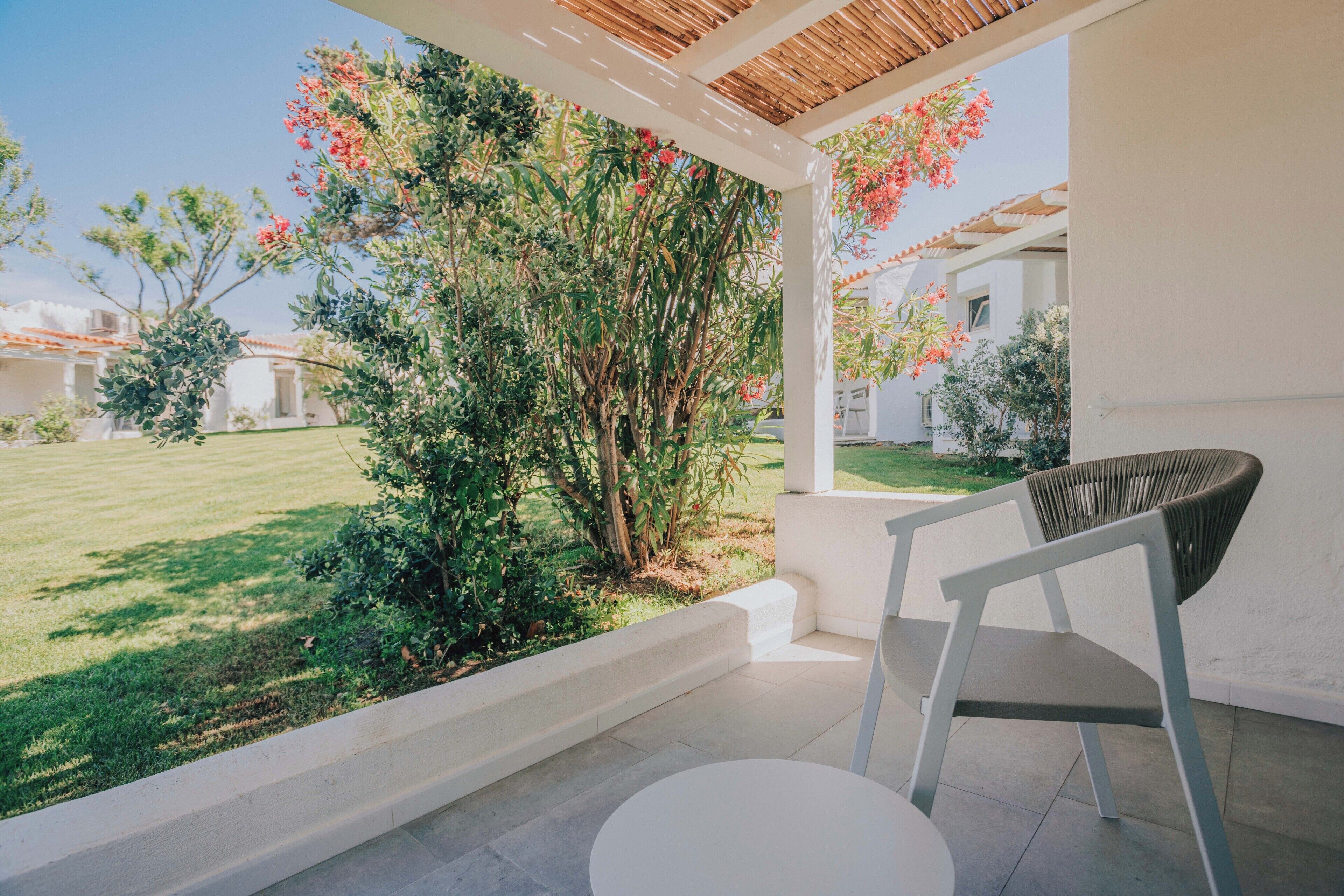 A covered patio with a white chair and table, overlooking a garden with red flowers and a lawn.