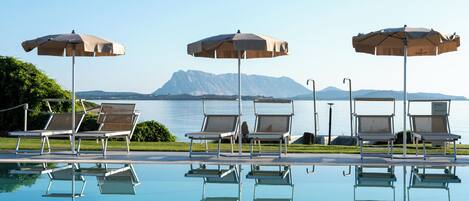 A poolside area with lounge chairs and umbrellas, a clear view of the water, and mountains in the background.