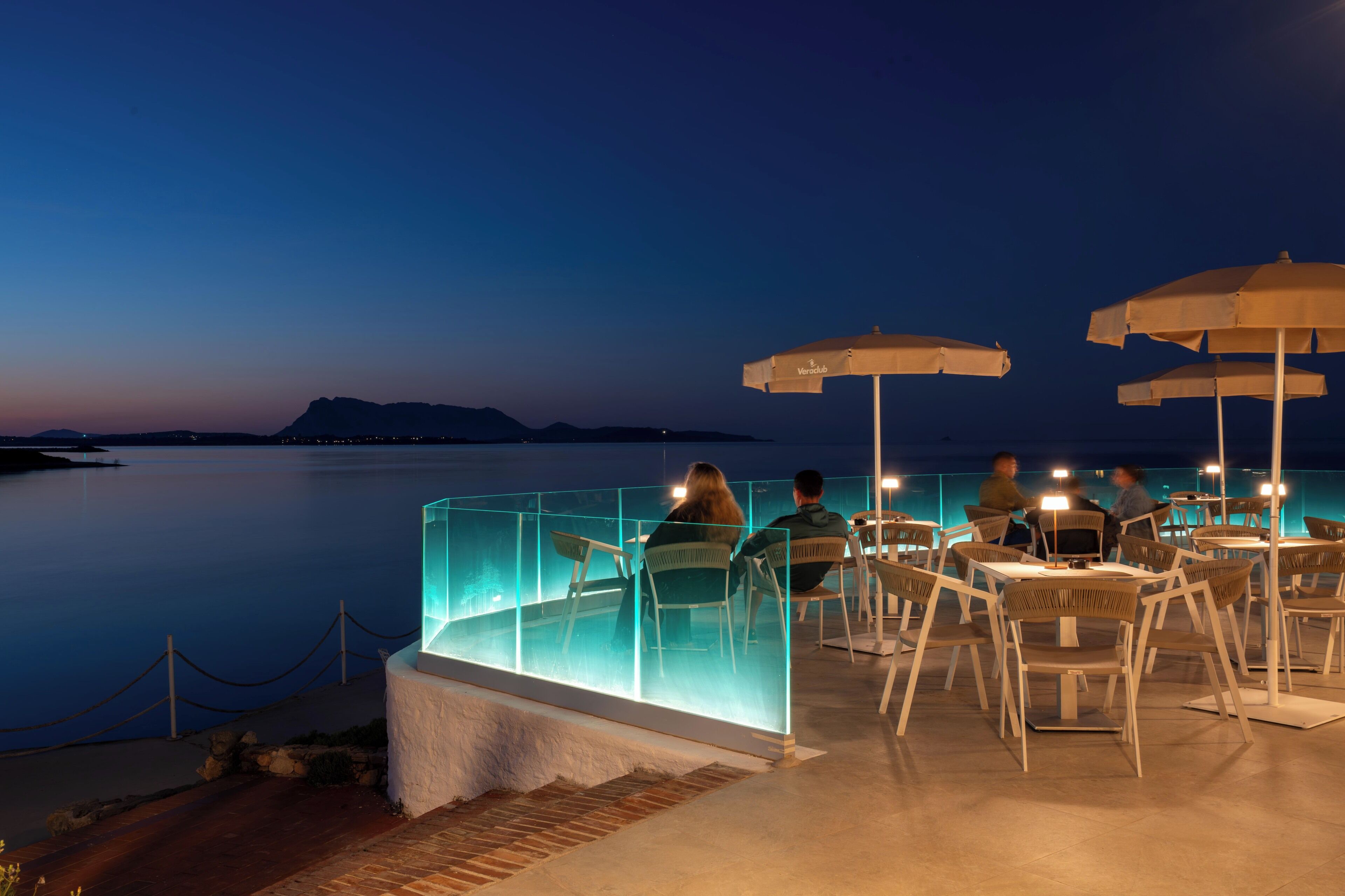 Un área de comedor al aire libre con mesas y sillas, con vista a una masa de agua tranquila al atardecer.
