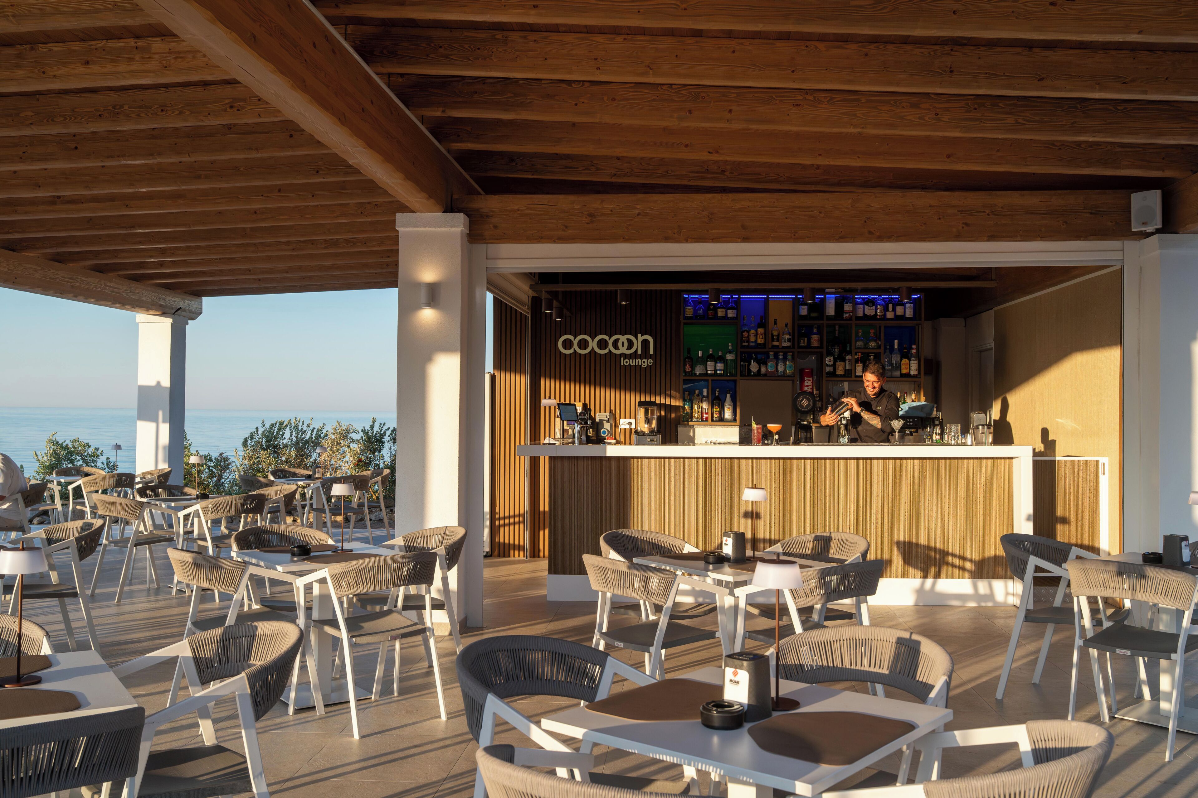 An outdoor seating area with wicker chairs and tables, a bar with a wooden counter, and a view of the sea.