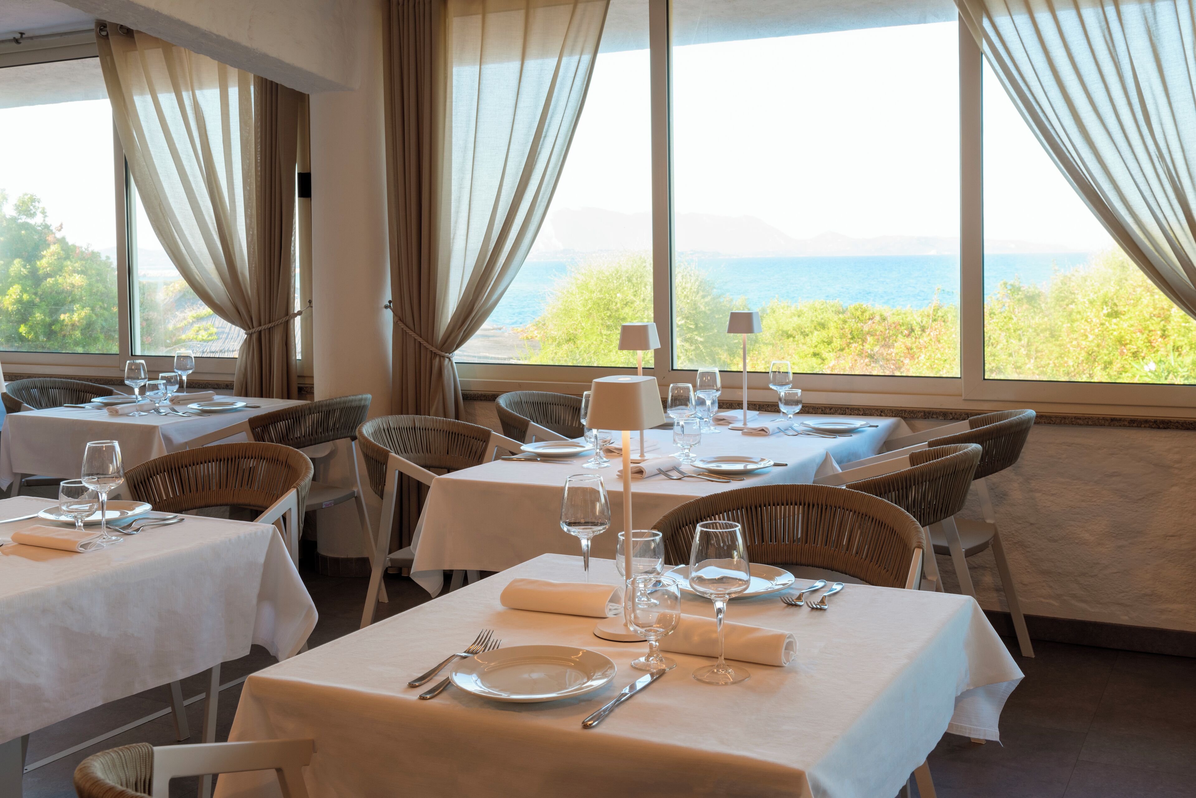 A restaurant with tables set for dining, large windows with curtains, and a view of the sea.